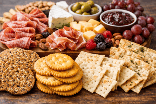 A charcuterie board featuring a variety of crackers including seeded, butter, and water crackers served with cheeses, cured meats, fruits, olives, and dips.
